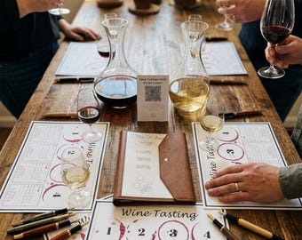 May include: A wine tasting set featuring a brown leather-bound journal titled The Wine Log, multiple printed tasting mats with numbered circles, two glass decanters, several wine glasses filled with red and white wine, and various fountain pens.