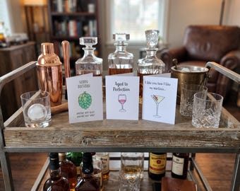 May include: Three white greeting cards displayed on a wooden bar cart. The left card features a green hop illustration with the text Hoppy Birthday Another Beer Best Another Beer Worst. The center card shows a wine glass with Aged to Perfection. The right card displays a martini glass with the text I like you how I like my martinis.