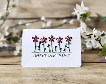 May include: A white greeting card with embroidered flowers and the text "HAPPY BIRTHDAY". The flowers are burgundy with green stems. The card is on a wooden surface with white flowers in the background.