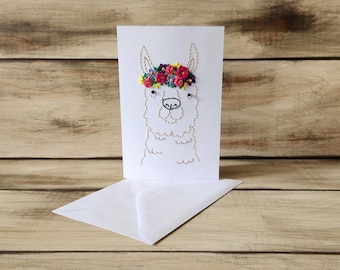 May include: A white greeting card featuring a hand-embroidered llama outline in tan thread. The llama wears a vibrant, three-dimensional floral crown made of pink, red, yellow, and blue embroidered flowers. A matching white envelope sits below.