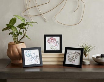 May include: Three square black frames containing line art prints of faces. One print features a blue butterfly, one a red rose, and one a green monstera leaf. The frames are arranged on a stack of books next to a potted plant and an air plant.