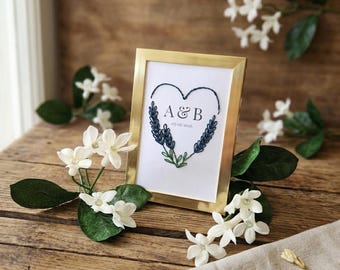 May include: A gold-toned rectangular frame containing a white insert with a dark blue embroidered heart shape. Inside the heart, the letters A & B are printed above the date 05-02-2026. The heart is formed by two sprigs of lavender.