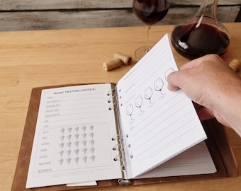 May include: A brown leather binder containing white paper sheets with printed wine tasting notes. The pages feature sections for name, date, and ratings using grape and wine glass icons. A fountain pen lies on the surface next to the open binder.