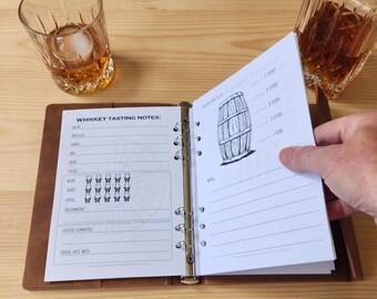 May include: A brown leather binder open to a whiskey tasting notes page with a barrel illustration, star ratings, and lines for entries. Beside it sits a glass of amber liquid, a glass decanter, and a wooden fountain pen on a light wood surface.