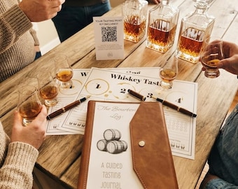 May include: A brown leather-bound tasting journal titled The Bourbon Log: A Guided Tasting Journal with a barrel illustration, accompanied by whiskey tasting mats, three glass decanters filled with amber liquid, and several small tasting glasses.