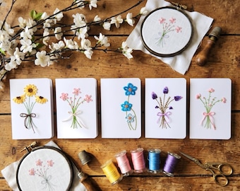 May include: Five white greeting cards featuring embroidered flower bouquets, two circular embroidery hoops with floral designs on white fabric, five spools of colored thread, a metal thimble, and gold scissors arranged on a wooden surface.