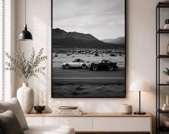May include: A large black-framed wall art print featuring a black and white photograph of two sports cars driving on a desert road with mountains in the background. The print is displayed above a wooden media console in a living room setting.