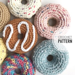 Op de afbeelding: Een verzameling kleurrijke, gehaakte donuts. De donuts zijn in verschillende kleuren, waaronder bruin, roze en blauw, met witte glazuur en hagelslag. De tekst "CROCHET PATTERN" is zichtbaar.