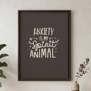 May include: A framed print with the text "ANXIETY is my SPIRIT ANIMAL" in white script and decorative elements on a dark gray background. The print is displayed above a wooden console table with a plant and decorative items.
