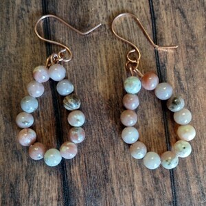 Puede incluir: Un par de pendientes en forma de lágrima. Cada pendiente presenta un bucle de pequeñas cuentas redondas multicolores en tonos rosa, gris y verde. Las cuentas están ensartadas en alambre de color cobre, con un gancho simple para la oreja.