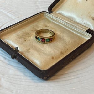 May include: A gold ring with alternating red and green gemstones, displayed inside a vintage brown jewelry box with a cream-colored satin lining. The ring is centered in the open box, showcasing its design.