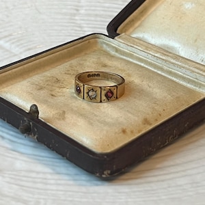 Op de afbeelding: Een gouden ring met drie ingezette edelstenen, twee rode en één witte, in een rechthoekig patroon. De ring wordt getoond in een bruine sieradendoos met een crèmekleurig interieur. De ring is waarschijnlijk vintage of antiek.