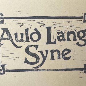May include: A vintage-style print on cream-colored paper. The text "Auld Lang Syne" is in dark blue, within a rectangular border with decorative corner elements. The font has a classic, slightly distressed appearance.