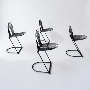 May include: Four black metal chairs with a minimalist design. Each chair features a curved backrest and seat made of thin, parallel metal rods. The legs have a unique, angular design with a semi-circular base, creating a modern aesthetic.
