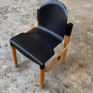 Può includere: Una sedia nera con schienale e seduta curvi, sostenuta da gambe in legno marrone chiaro. La sedia ha un design moderno con un'estetica minimalista. Le gambe sono cilindriche e lo schienale è arrotondato.