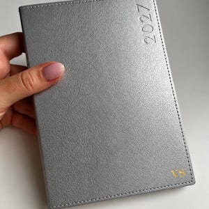 May include: A rectangular silver planner with a textured finish, featuring the year 2027 embossed vertically along the right edge and the gold-coloured initials VS stamped in the bottom right corner, with visible stitching along the outer border.