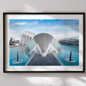 May include: Framed print featuring a modern architectural complex with a large, curved building and other structures reflected in the turquoise water. The sky is blue with a few clouds. The print is in a black frame.