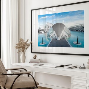 May include: A framed photograph of a modern architectural structure with a blue sky and water. The building has a unique, curved design with glass and white elements. The photograph is displayed on a white desk with a chair, vase, and other decorative items.
