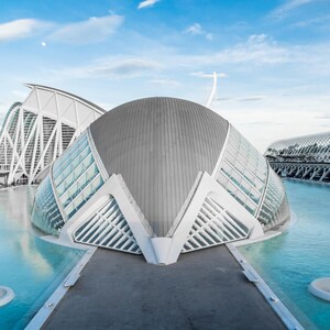 May include: Architectural photograph of the City of Arts and Sciences in Valencia, Spain. The image features modern, white, and gray structures with glass panels, set against a blue sky and water. The buildings have unique, curved designs.
