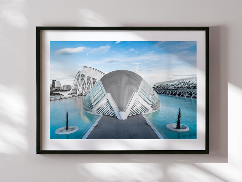 May include: Framed print featuring a modern architectural complex with a large, curved building and other structures reflected in the turquoise water. The sky is blue with a few clouds. The print is in a black frame.