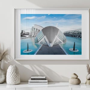 May include: Framed print featuring a modern architectural complex with a futuristic design. The buildings have white, curved structures and are reflected in the turquoise water. The sky is blue with a few clouds. The print is in a white frame.