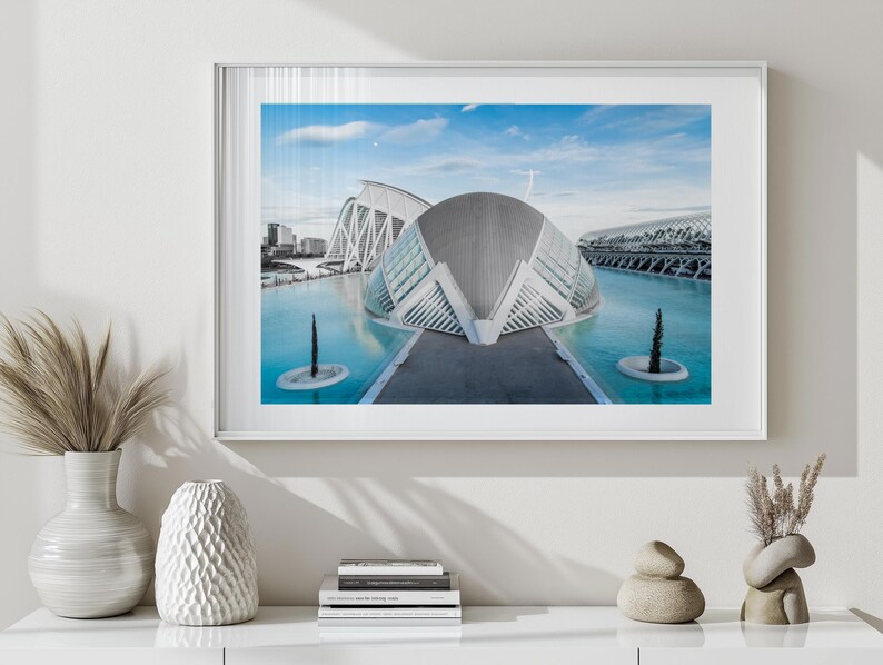 May include: Framed print featuring a modern architectural complex with a futuristic design. The buildings have white, curved structures and are reflected in the turquoise water. The sky is blue with a few clouds. The print is in a white frame.