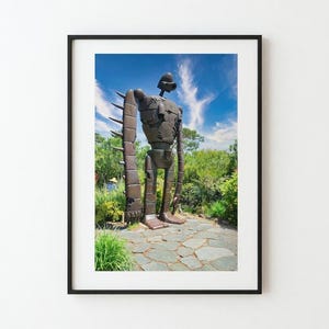 Puede incluir: Impresión enmarcada de una gran estatua de robot de color bronce con un casco, de pie al aire libre. El robot tiene un cuerpo rectangular y brazos largos con púas. El fondo presenta un cielo azul con nubes y vegetación exuberante.