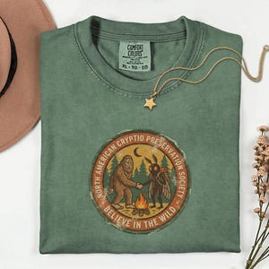 Può includere: T-shirt verde Comfort Colors con una grafica rotonda raffigurante Bigfoot e un Mothman che si stringono la mano. La grafica recita "North American Cryptid Preservation Society, Believe in the Wild". Una collana con pendente a stella dorata è appoggiata sulla maglietta.