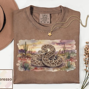 Op de afbeelding: Espresso-kleurig T-shirt met een aquarel illustratie van een opgerolde ratelslang in een woestijnlandschap met cactussen. Het shirt heeft een ronde hals en een Comfort Colors label. Een gouden ketting met een sterhanger is over het shirt gedrapeerd.