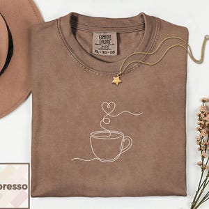 May include: Espresso-coloured t-shirt featuring a white line drawing of a coffee cup and steam forming a heart shape. A gold star pendant necklace rests on the shirt. The shirt has a Comfort Colors tag.