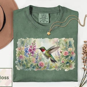 May include: Green t-shirt with a watercolor hummingbird design. The shirt features a vibrant hummingbird with red, green, and white feathers, hovering near colorful flowers. A gold necklace with a star pendant is on the shirt. The tag reads "Comfort Colors XL-TG-EG".