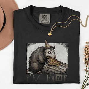 Könnte beinhalten: Schwarzes Comfort Colors T-Shirt mit einer verwitterten Illustration eines Opossums, das eine Partyhütchen trägt und Pizza aus einem Mülleimer isst. Der Text "I'M FINE" ist darunter gedruckt. Eine goldene Sternanhänger-Halskette liegt auf dem Shirt.