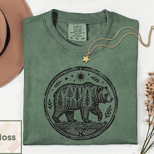 May include: Moss green t-shirt with a black bear graphic inside a circular design. The bear's fur is made of trees. The shirt has a crew neck and a Comfort Colors tag. A gold star pendant necklace is on the shirt.