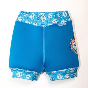 Può includere: Pantaloncini da nuoto blu brillante con fascia in vita e bordi delle gambe decorati con una stampa di bolle azzurre e facce da cartone animato. Una piccola etichetta bianca con il testo Swim Nappy è attaccata al lato. Il pantaloncino ha un pannello centrale blu.