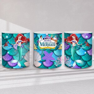 May include: White ceramic mug featuring Ariel from Disney's The Little Mermaid. The mug has a teal, purple, and blue scale pattern background. Ariel is depicted with red hair and a green tail. The mug has a white handle.