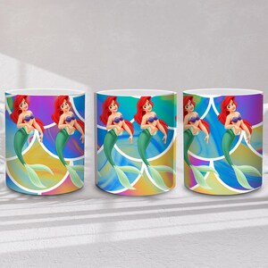 May include: Three white ceramic mugs featuring a mermaid with red hair, a purple seashell bra, and a green tail. The mermaid is set against a colorful, abstract background. Each mug has a white handle.
