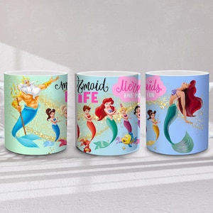 May include: Three white ceramic mugs with mermaid-themed designs. One mug features King Triton, another displays mermaids with the text "Mermaid Life", and the third showcases a mermaid with red hair. Each mug has a white handle.