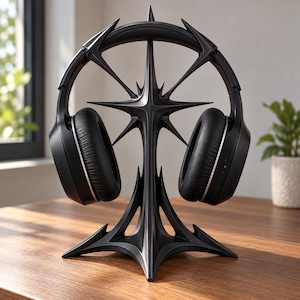 Puede incluir: Un soporte para auriculares negro con un diseño de estrella de múltiples puntas afiladas, que presenta un pilar central vertical y una base ancha y estable. Unos auriculares negros con almohadillas descansan sobre el soporte.