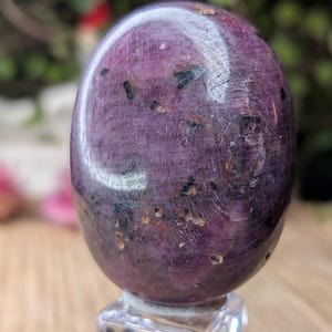 Può includere: Una pietra di rubino zoisite lucidata di forma ovale con una base color magenta e viola intenso, caratterizzata da inclusioni di orneblenda nera sparse e piccole macchie marrone dorato su tutta la superficie, poggiata su un supporto quadrato in acrilico.