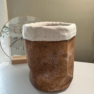 May include: A cylindrical, handmade ceramic planter with a textured, brown exterior and a thick, white-painted rim. The planter has a rustic, organic aesthetic, suitable for indoor or outdoor use. The planter is designed to hold plants.
