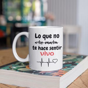 May include: White ceramic mug with a curved handle. The mug features black text that reads "Lo que no te mata te hace sentir vivo" with a heartbeat line and heart symbol. The mug is on a book.