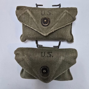 Puede incluir: Dos bolsas de lona verde oliva con cierres metálicos y las letras "U.S." impresas. Las bolsas son rectangulares con cierre de solapa y están montadas sobre soportes metálicos negros. La lona tiene un aspecto texturizado.