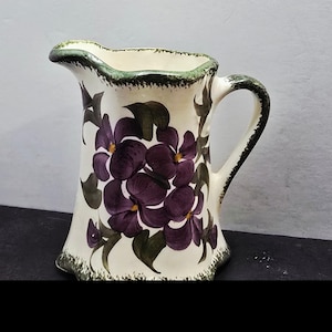 May include: A cream-colored ceramic pitcher featuring a hand-painted floral pattern of deep purple flowers with yellow centers and dark green leaves. The rim and handle are accented with a textured, dark green sponged edge detail.