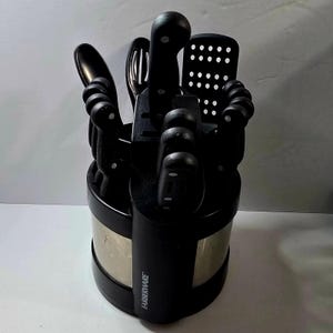 May include: A black Farberware knife block set containing various kitchen utensils. The set includes knives, a slotted spoon, and a spatula. The knife block has a black exterior with a silver band. The utensils are arranged in a circular pattern.