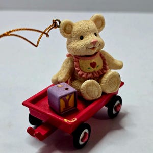 Puede incluir: Adorno decorativo con un osito de peluche beige con un babero con un corazón, sentado en un vagón rojo. Un bloque morado con la letra "Y" está en el vagón. El adorno tiene una cuerda dorada para colgarlo.