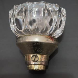 May include: A clear, faceted glass doorknob with a silver-colored metal base and a brass-colored metal stem. The glass knob has a rounded, floral-like design. The doorknob is likely vintage or antique.