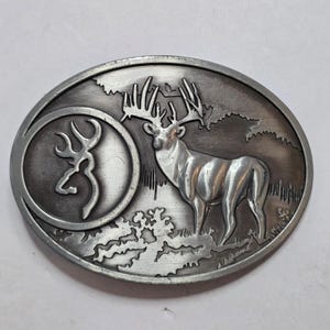 May include: Oval-shaped silver-tone belt buckle featuring a detailed depiction of a buck with large antlers in a forest scene. The left side has a circular emblem with a deer silhouette. The buckle is likely made of metal.