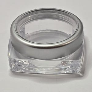 May include: A clear, square cosmetic jar with a silver-colored lid. The jar is made of transparent plastic, allowing visibility of the contents. The lid has a smooth, metallic finish. The jar is designed for storing beauty products.