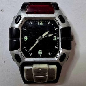 Op de afbeelding: Een zilver en zwart polshorloge met een zwarte wijzerplaat, witte wijzers en cijfers. Het horloge heeft knoppen met de tekst "POWER", "CABLE" en "MUTE/SET". Een rood rechthoekig element bevindt zich bovenaan.
