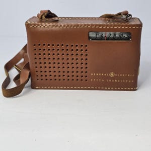 May include: A vintage brown leather General Electric Seven Transistor radio with a carrying strap. The radio has a speaker grill with multiple holes and a tuning dial. The text "GENERAL ELECTRIC SEVEN TRANSISTOR" is printed on the front.